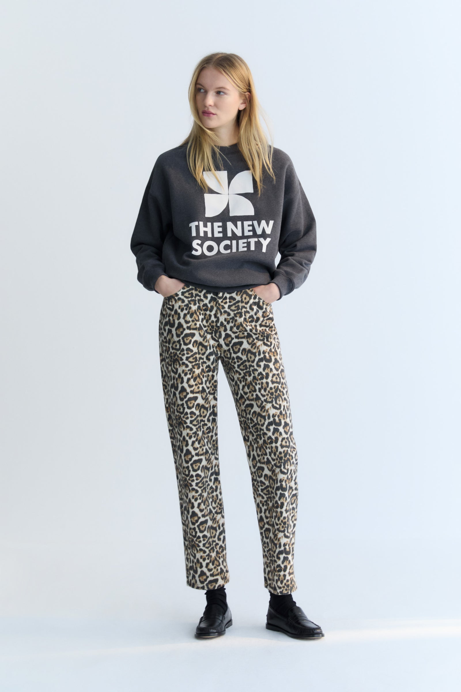 Person wearing a black sweatshirt with 'The New Society' logo and leopard print pants on a light gray background