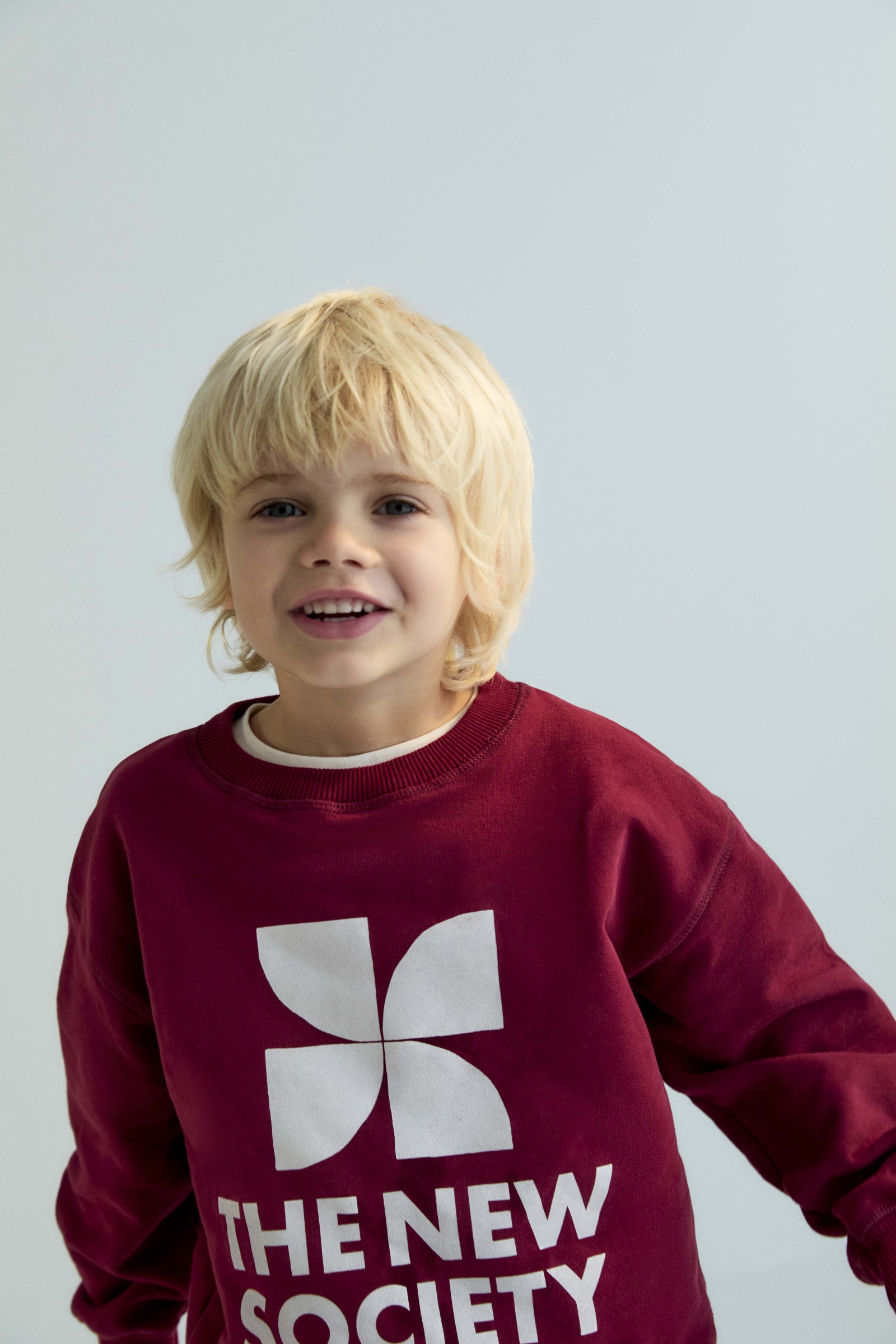 Child wearing a maroon sweatshirt with 'The New Society' logo on a light gray background